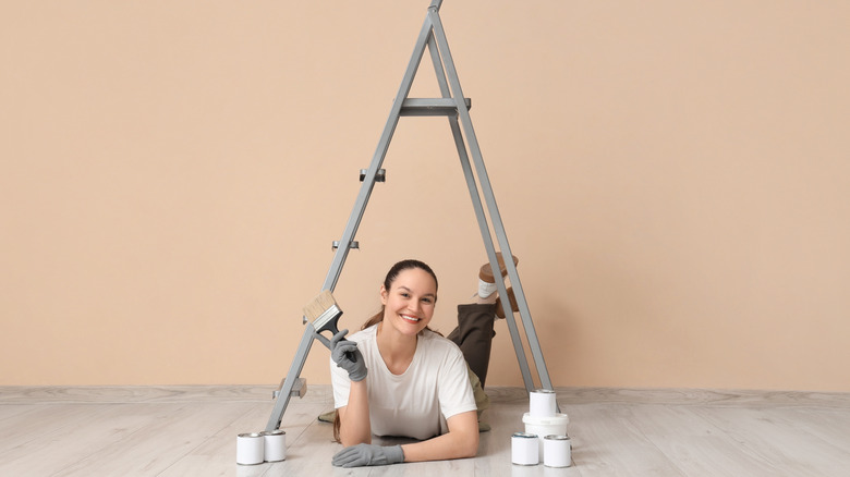 A smiling young woman with a brush and paint cans lying under stepladder after painting the wall beige.