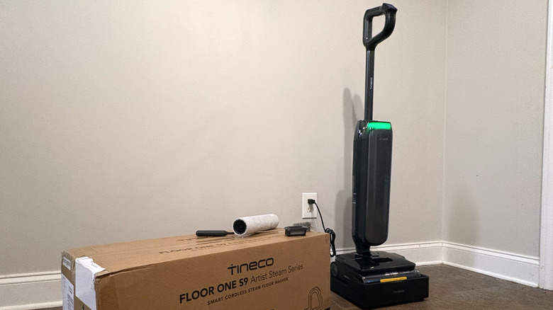 A Tineco Floor One S9 Artist Steam Series Smart Cordless Steam Floor Washer sits in a charging station next to its accessories and packaging