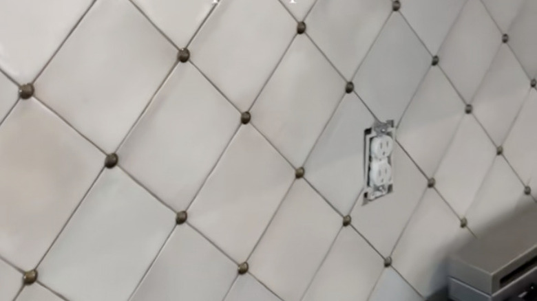 A stretch of ceramic tile wall is dotted with brass thumbtacks between the grouted joints.