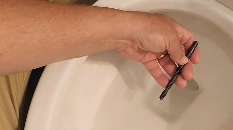 Man's hand holding a drill bit in toilet bowl, preparing to use it to clean the siphon jet.