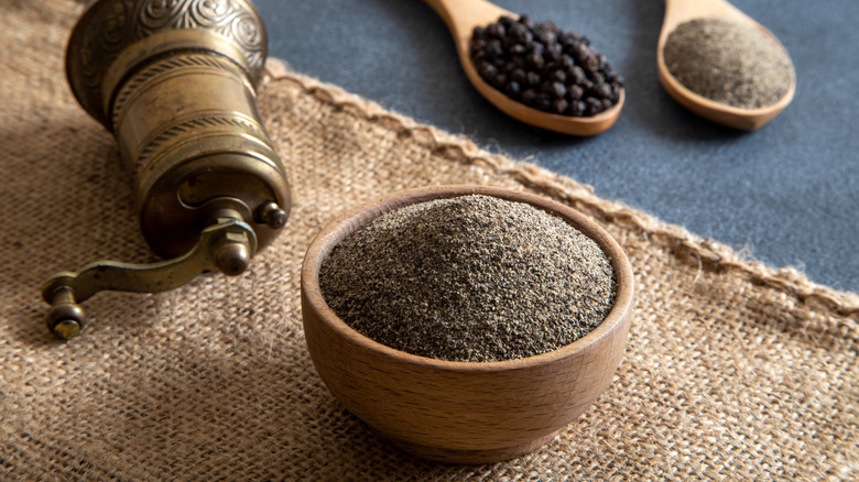 Wooden bowl of ground pepper sits on a woven mat next to a pepper grinder and spoons of whole peppercorns