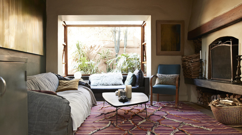 A living room with a big open window, a fireplace, a grey couch, a coffee table, and a woven mauve rug.