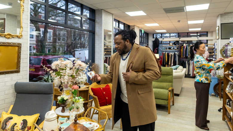A man observes a tea cup in a thrift store while a woman looks at d&eacute;cor on nearby shelves.