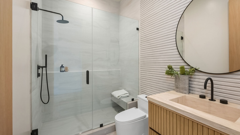 Modern bathroom interior with a glass-enclosed shower, wooden vanity, and large round mirror, featuring minimalist design elements.
