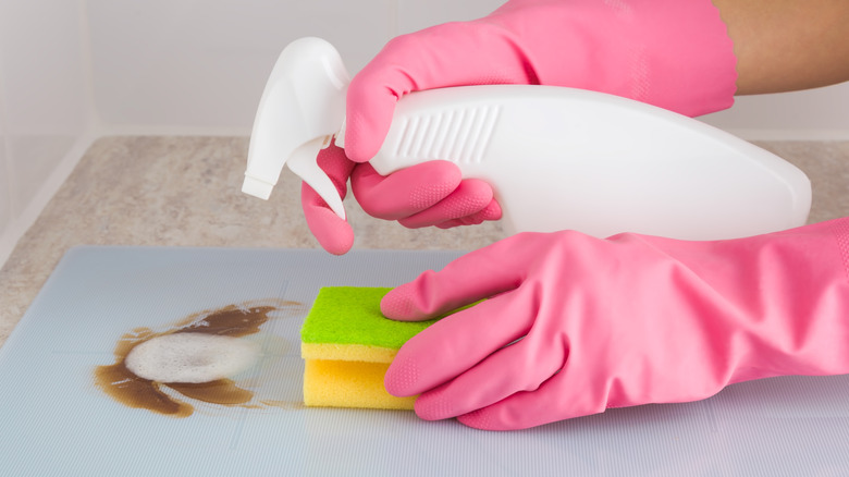 Hands in pink rubber gloves holding a white spray bottle aimed at a stovetop grease stain