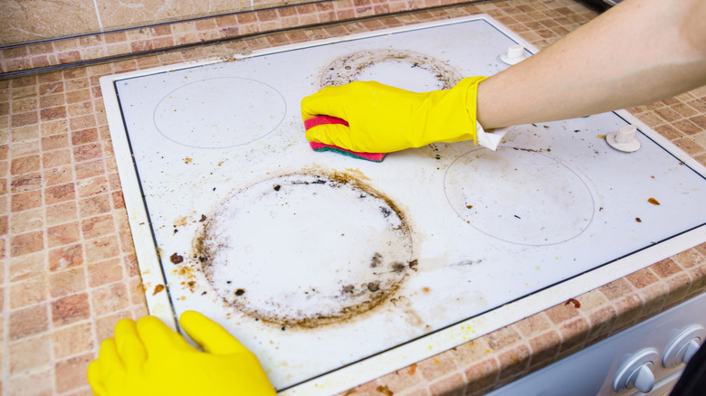 Hands in yellow rubber gloves using a sponge to clean a greasy, dirty electric stove top