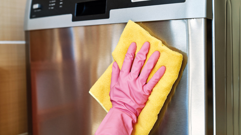 Gloved hand wiping a stainless steel dishwasher with yellow cloth