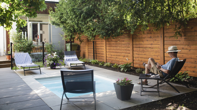 A man reading a book while sitting near a backyard pool surrounded by a wood fence