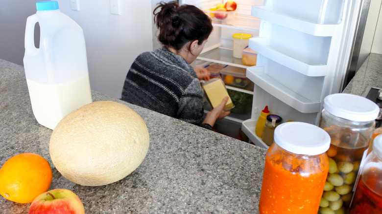 A woman sits on the floor to clean her refrigerator. Food items can be seen scattered on the counter behind her