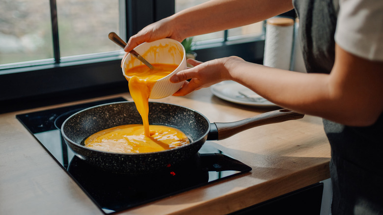 Person in apron pours scrambled eggs out of a bowl into a pan on a cooktop.