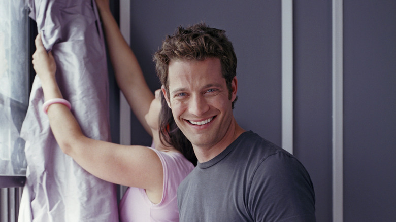 Nate Berkus smiling while a woman hangs a curtain in the back