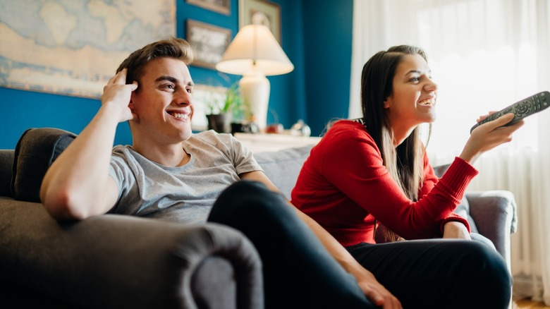 A man and a woman watching TV while the woman holds the tv remote