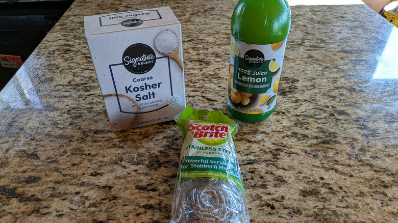 A box of kosher salt, a bottle of lemon juice, and a pack of metal scrubbers sit on a kitchen countertop.