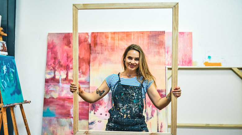 Woman in paint covered overalls holding a half-built picture frame with canvas painting behind her