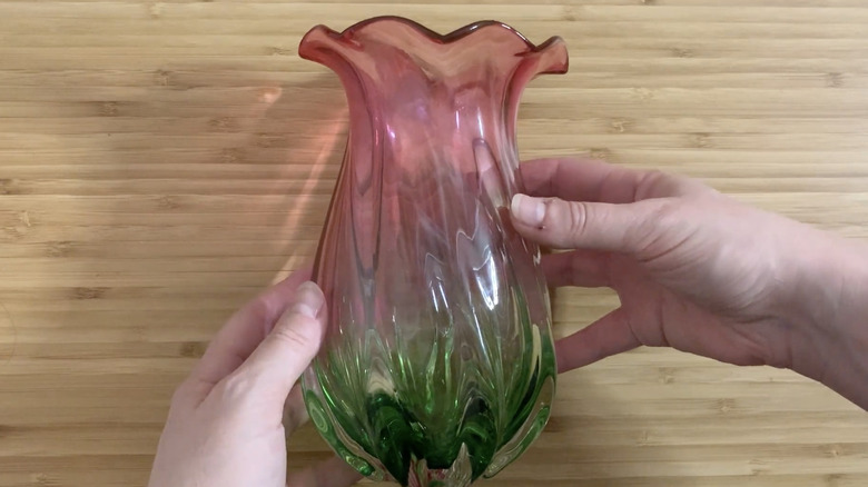 A pair of hands holding the pink and green watermelon-style Teleflora vase above a wood surface.