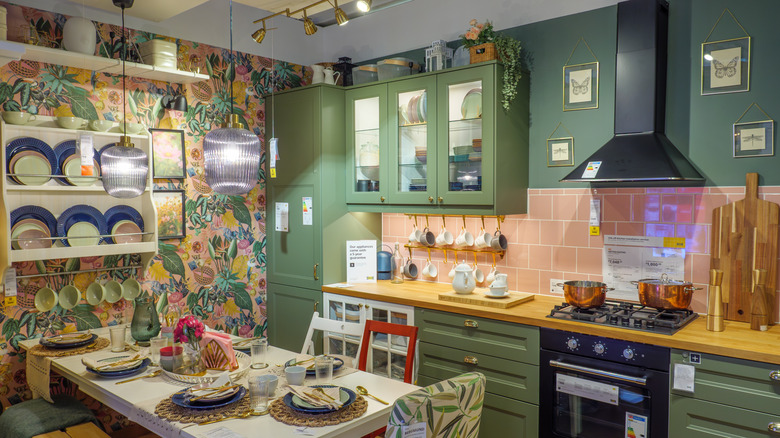 IKEA store display of kitchen featuring items for sale. Green cabinets, pink tile backsplash, and botanical patterned wallpaper.