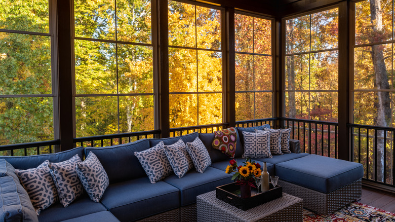A three season porch with large windows and sectional seating surrounded by leafy trees through the windows