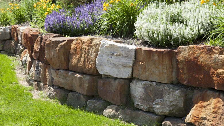 A brown stone retaining wall holds back soil with flower beds and plants.