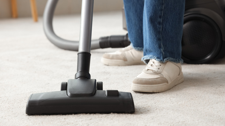 Close up of vacuum head and a person's feet and lower legs vacuuming a carpet.