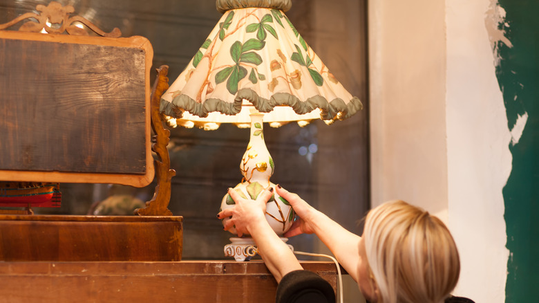 A woman reaches up to pick an old floral lamp from a shelf.