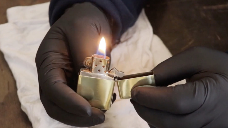 A man wearing black gloves holds the lit metal lighter he just restored over a white rag on a tabletop.