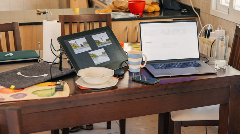 A cluttered wood kitchen table with a bowl, cup, laptop, and tablet