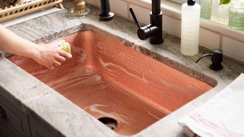 A hand wiping a copper sink with a soapy sponge