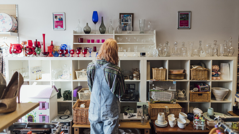 A woman stands in a thrift store admiring all the glassware arranged nicely on the display shelves.