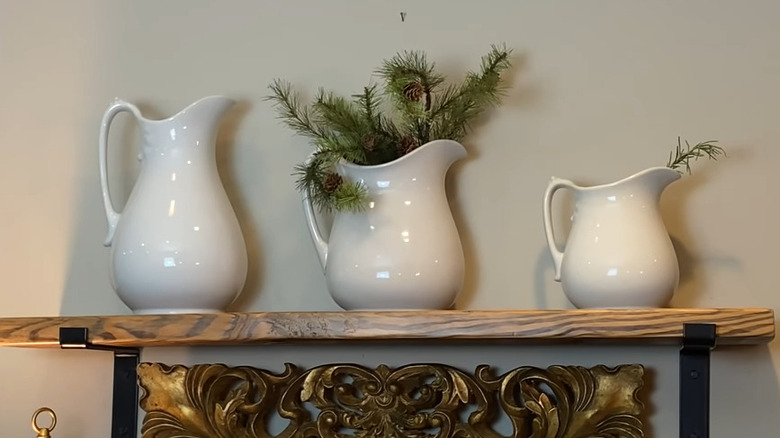 Three ironstone pitchers, two holding sprigs of pine and rosemary, on a wooden shelf