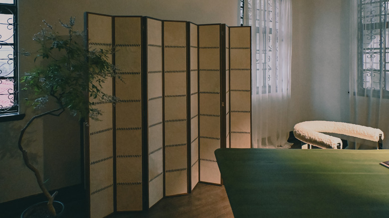 A multi-panel wooden folding screen stands in front of a wall with a tree and furniture on either side.