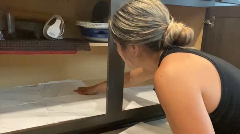 A person installing vinyl floor tiles in their kitchen cabinets