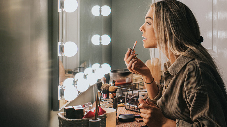 Woman putting on makeup at cluttered bathroom mirror