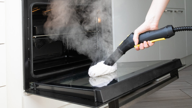 A hand holding a steam cleaner on the glass inside an oven with steam billowing up.