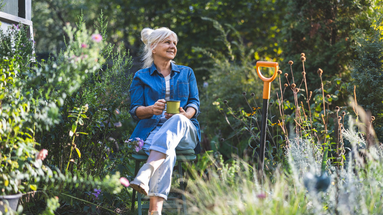 A person sitting in the garden with a hot beverage next to a yellow-handled garden tool