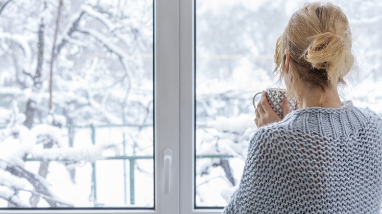 Woman in gray sweater holding mug look outside at snow