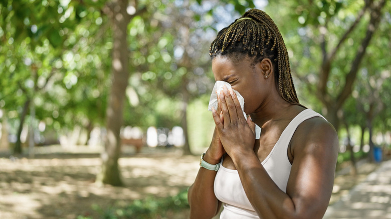 A young woman with blows her nose under a leafy tree.