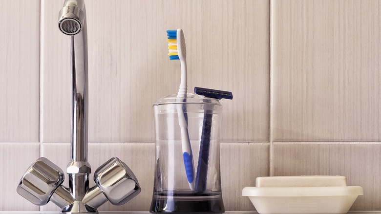 A water tap, toothbrush and razor in a cup, and a bar of soap in a soap dish in a bathroom