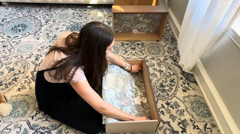 A woman is lining a drawer with a cut piece of wallpaper