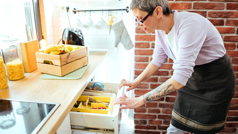 A person wearing an apron in the kitchen opening a drawer with utensils