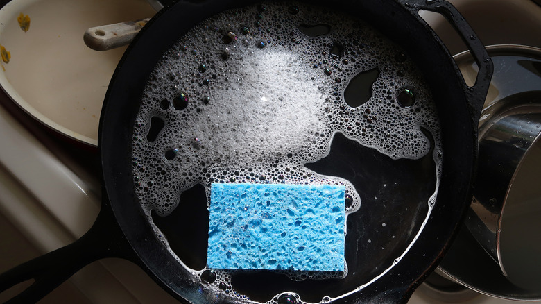 Cast iron frying pan in dirty sink with sponge and soapy water