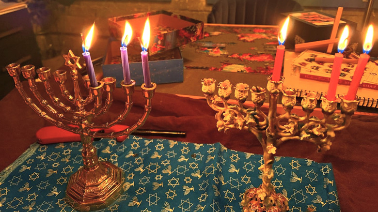 Two menorahs with intricate design and burning candles placed atop a table with other Hanukkah decor.