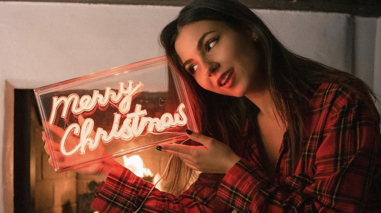 Victoria Justice holding a Merry Christmas neon sign close to her face while sitting by the fireplace and wearing plaid pajamas.