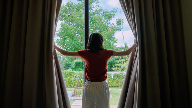 View of woman opening blackout curtains to a bright day, backyard with trees and hedges.