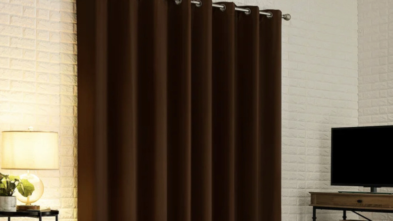 Product image of a brown Sun Zero Broderick black out curtains displayed over a window. Living room, TV on a stand and a nightstand with a table lamp on top.