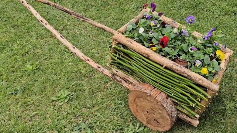 A person fastens a wooden wheel to an axle and a wheelbarrow made of branches planted with colorful flowers on a lawn.