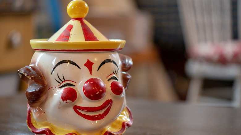 A cookie jar in the shape of a clown with a tent top for the lid sitting on a wooden table top.