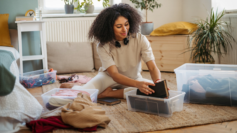 Young woman sorting books and bins of clothes on floor of bedroom