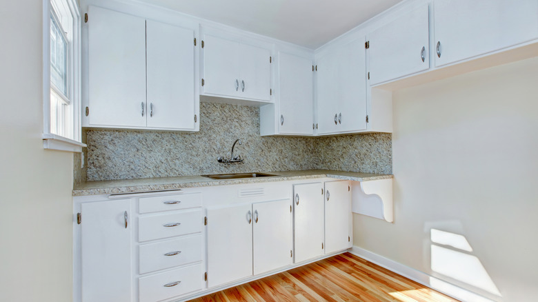 A small corner kitchen with white cabinets, a granite backsplash with matching countertops, and a wooden floor.