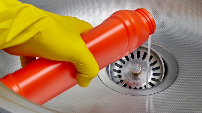 Person wearing a yellow glove while pouring a drain cleaner chemical out of a red bottle into a sink drain