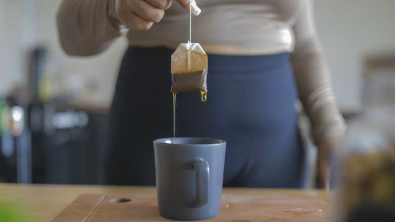 A woman standing behind a blue mug pulling a dripping tea bag out of the mug with one hand